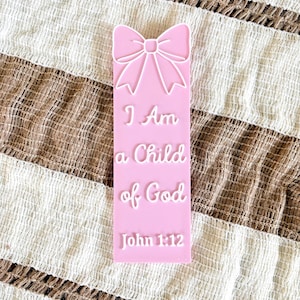 May include: A pink bookmark with a bow design at the top and the text "I Am a Child of God John 1:12" in white. The bookmark is rectangular and has a raised design. The background is a striped, textured fabric.