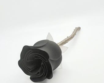 May include: A single black rose with a detailed, layered petal design. The stem is a light brown color, and the leaves are a translucent white. The rose is set against a plain white background.