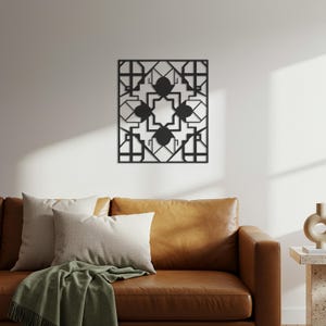 May include: A black metal wall art piece with a geometric design, featuring squares, lines, and star-like shapes. The artwork is mounted on a white wall above a brown leather sofa with pillows and a green blanket.