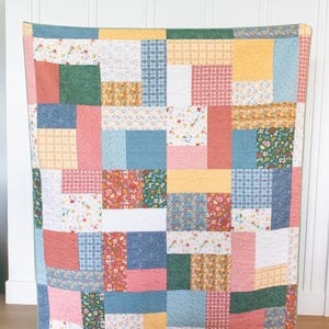 May include: A patchwork quilt with a variety of fabric patterns and colours. The quilt features squares of floral, plaid, and solid fabrics in shades of pink, blue, green, yellow, and white. The quilt is displayed against a white wall.