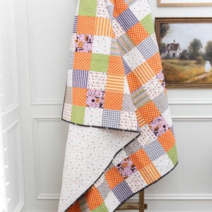 Puede incluir: Un edredón de retazos con temática de Halloween, colgado de una escalera de madera. El edredón presenta cuadrados de telas naranjas, verdes, moradas y blancas. Algunos cuadrados tienen patrones temáticos de Halloween. El borde del edredón es negro.