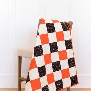 Puede incluir: Un edredón hecho a mano sobre una silla. El edredón presenta un patrón de tablero de ajedrez de cuadrados naranjas, negros y crema. Algunos cuadrados negros tienen un diseño floral. El edredón tiene un borde naranja.