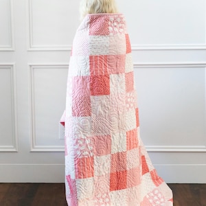 Puede incluir: Un edredón hecho a mano con un diseño de patchwork en tonos rosa y blanco. El edredón presenta varios patrones dentro de los cuadrados, incluidos lunares y motivos florales. El edredón está cubriendo a una persona invisible.