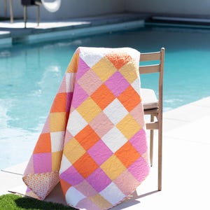 Può includere: Una colorata trapunta con un motivo geometrico di quadrati arancioni, rosa, gialli e bianchi è drappeggiata su una sedia di legno. La trapunta è vicino a una piscina e a un pezzo di erba sintetica verde.