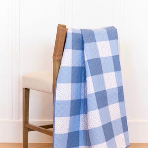 Puede incluir: Un edredón a cuadros azules y blancos sobre una silla de madera. El edredón presenta cuadrados grandes de diferentes tonos de azul y blanco, con un patrón floral más pequeño en algunos cuadrados. La silla tiene un asiento beige claro y un respaldo de cuero marrón.