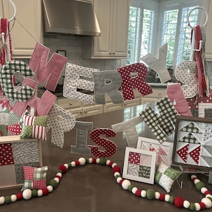 May include: A festive Christmas display featuring a "MERRY IS" banner with fabric letters in red, green, and gray patterns. Framed fabric art and a garland with red, white, and green beads complete the holiday theme.