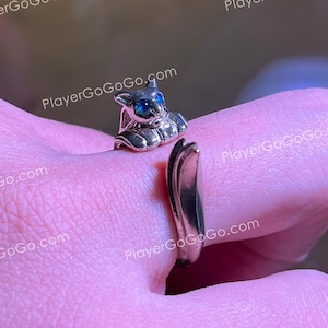 May include: A gold-colored ring featuring a small cat figurine with blue eyes. The ring is wrapped around a finger, showcasing the cat's detailed design. The ring is a piece of jewelry.