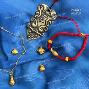 May include: A gold-toned necklace with a small bottle pendant, a red braided cord bracelet with a gold-toned fish charm, a brass-colored decorative plaque with engraved Chinese characters, and three small gold-toned charms on a blue fabric.