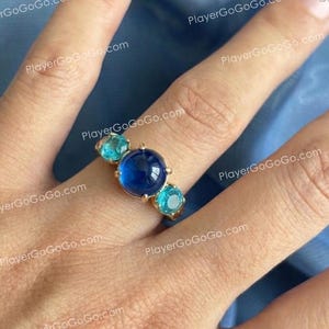 Pode incluir: Um anel de ouro com uma pedra cabochão grande, redonda e azul-escura no centro, ladeada por duas pedras redondas mais pequenas de cor turquesa brilhante em cada lado. O anel é montado numa banda de ouro polido com cravação de garras.