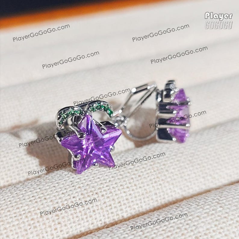 May include: A pair of silver earrings featuring a purple star-shaped gemstone. The earrings have a small green accent and a hinged clasp. The earrings are displayed on a textured, off-white surface.