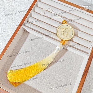 May include: A gold-colored pendant with Chinese characters, attached to a white and yellow tassel. The pendant hangs inside a wooden jewelry box with a light-colored fabric lining.