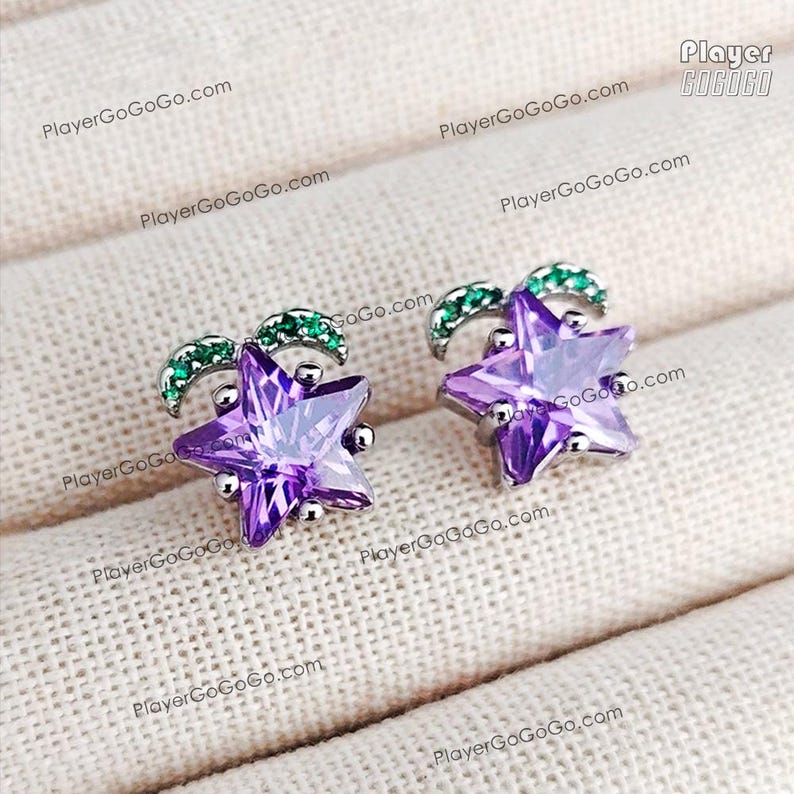 May include: A pair of star-shaped stud earrings. Each earring features a faceted purple gemstone star, accented with a green, curved design at the top. The earrings are set in silver-colored metal and rest on a textured, off-white surface.