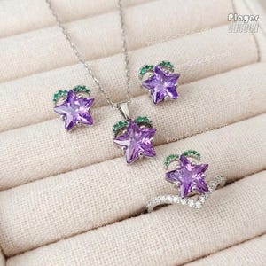 May include: A jewelry set consisting of a silver necklace, two stud earrings, and a ring. Each piece features a purple star-shaped gemstone accented with small green stones at the top. The ring has a V-shaped band set with clear stones.