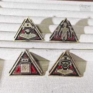 May include: Four triangular metal pins with various designs. Each pin features a silver-colored frame with a red and black interior. The designs include a figure wearing a helmet, a group of figures, and a rectangular object. The pins are displayed on a textured surface.