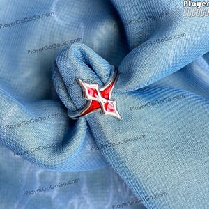 Puede incluir: Un anillo plateado con un diseño geométrico rojo y blanco. El anillo presenta una forma roja central con detalles blancos, engastada en una banda plateada. El anillo se muestra sobre una tela azul.