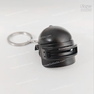 May include: A dark gray helmet keychain with a silver key ring. The helmet has a visor and is likely a miniature replica of a military or gaming helmet. The text "PlayerGoGoGo.com" is visible.