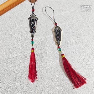 May include: Two decorative pendants with black and gold filigree designs, red accents, and long red tassels. Each pendant has a string of beads in green, red, and clear colors. The pendants are hanging against a textured white background.