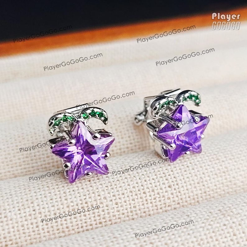 May include: A pair of silver-toned star-shaped stud earrings. Each earring features a faceted purple gemstone star and a curved green accent. The earrings are displayed on a textured, off-white surface.