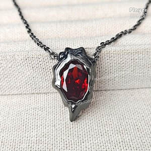 Può includere: Una collana in metallo nero con un ciondolo con una grande pietra preziosa rossa sfaccettata. Il ciondolo ha una forma irregolare e organica, e la catena è di un colore scuro abbinato. La gemma è di un rosso intenso e vibrante.