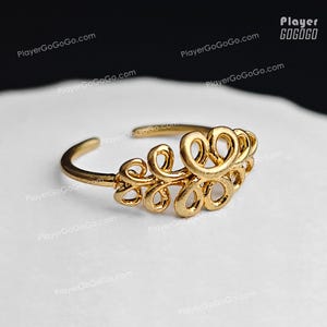 May include: A gold-colored ring with an open design. The ring features an ornate, swirling pattern across the top, with a simple band. The ring is set against a white background.