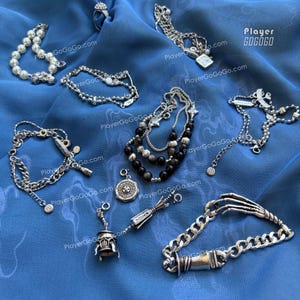 Op de afbeelding: Een collectie zilverkleurige sieraden, waaronder een parelarmband, een kettingarmband met een skelet-handsluiting, een meerstrengs kralenarmband en diverse bedels met een bel, een kompas en een ring, allemaal voorzien van het label PlayerGoGoGo.
