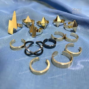 Może przedstawiać: Kolekcja złotych i czarnych pierścionków, w tym regulowane opaski i geometryczne wzory. Niektóre pierścionki mają kształt błyskawic i diamentów. Pierścionki są prezentowane na niebieskim tle z tkaniny.