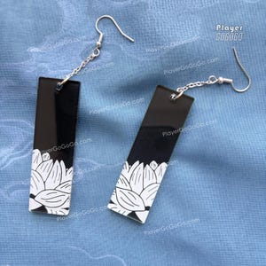 Puede incluir: Un par de pendientes rectangulares negros con un diseño floral blanco en la parte inferior. Los pendientes están conectados a ganchos plateados con una pequeña cadena. El fondo es de tela azul claro.