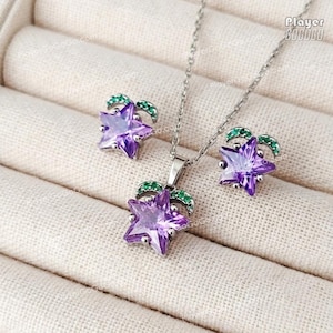 May include: A silver-toned jewelry set featuring a pendant necklace and matching earrings. Each piece showcases a faceted purple star design, accented with green gemstone details. The set is displayed on a neutral, textured surface.
