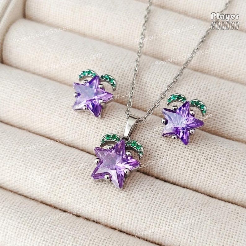 May include: A silver-toned jewelry set featuring a pendant necklace and matching earrings. Each piece showcases a faceted purple star design, accented with green gemstone details. The set is displayed on a neutral, textured surface.