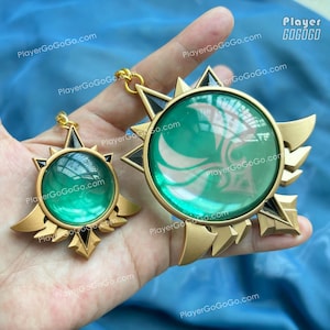 May include: Two gold-coloured pendants with a sunburst design. Each pendant features a large, round, emerald-green centre with a white design. The pendants have gold-coloured accents and black triangular details. One pendant is larger than the other.