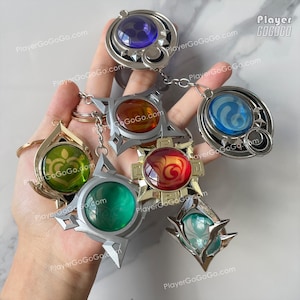Puede incluir: Una colección de siete llaveros metálicos con centros de colores similares a piedras preciosas. Cada llavero tiene un diseño único, incluyendo formas circulares, de estrella y de lágrima. Los llaveros presentan el texto "PlayerGoGoGo.com".