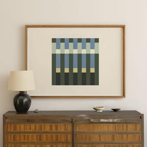May include: A framed abstract art print featuring a geometric design with vertical and horizontal lines in shades of green, blue, and yellow. The artwork is displayed in a wooden frame and hangs on a neutral-colored wall. A lamp and a wooden cabinet are in the foreground.