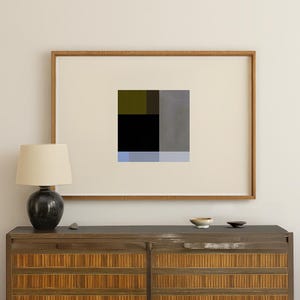 Puede incluir: Impresión de arte abstracto en un marco de madera. La obra de arte presenta un diseño geométrico con bloques de negro, gris, verde oliva y azul. La impresión se muestra encima de un mueble de madera con una lámpara y cuencos pequeños.