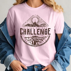 May include: A light pink t-shirt with a brown graphic design. The design includes the text "RISING TO THE CHALLENGE" and other phrases like "LAID MY FOUNDATION" and "ADDED THE ESSENTIALS". The graphic also features a mountain and forest scene.