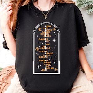 May include: Black t-shirt featuring a white and gold graphic with a crescent moon, stars, and the names of characters from a book series. The names are arranged in a vertical list within a rectangular frame. The shirt is made of a soft material.