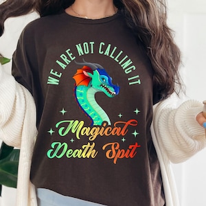 May include: A dark brown t-shirt with the text "WE ARE NOT CALLING IT" and "Magical Death Spit" in a whimsical font. The shirt features a colourful dragon graphic with blue, green, and red accents. The shirt is paired with light blue shorts.