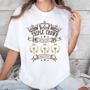 May include: White t-shirt featuring a graphic design with three crowns, the words "Triple Crown Memory Master", and gold laurel wreaths. The shirt is a casual, unisex style.