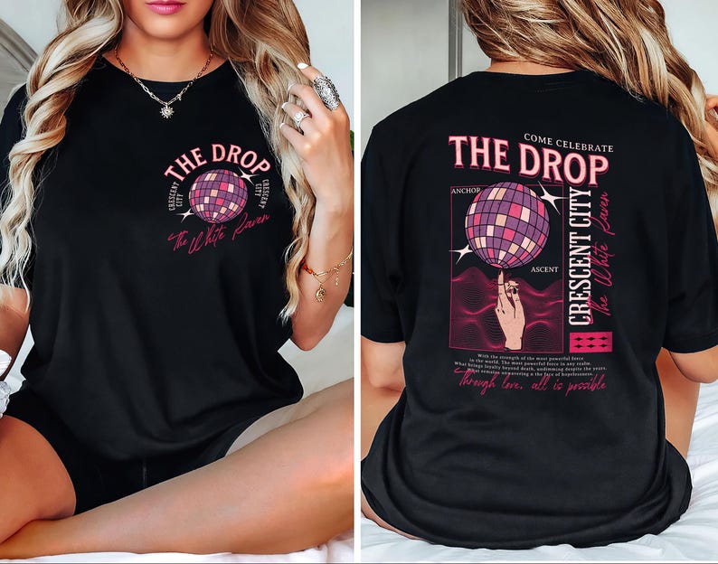 May include: Black t-shirt with a graphic design featuring a disco ball and the text "THE DROP" in pink. The design also includes the words "CRESCENT CITY" and "The White Raven". The back of the shirt has the same design.