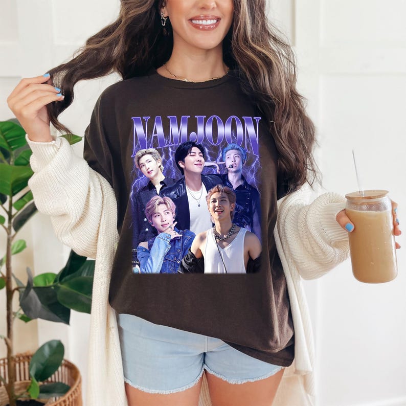 May include: A brown t-shirt featuring a graphic of a group of people with the word "NAMJOON" in a purple font. The graphic has a vintage style with a lightning bolt effect. The shirt is worn with light blue shorts and a white cardigan.