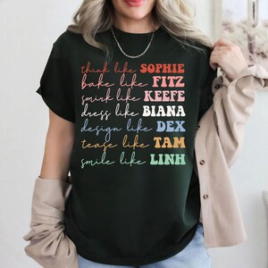 Puede incluir: Camiseta verde oscuro con texto colorido. La camiseta presenta las frases "think like SOPHIE", "bake like FITZ", "smirk like KEEFE", "dress like BIANA", "design like DEX", "tease like TAM" y "smile like LINH".