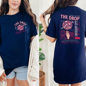 May include: Navy blue t-shirt with a graphic of a disco ball and the text "The Drop". The back of the shirt features a similar design with additional text including "Crescent City" and "Anchor".