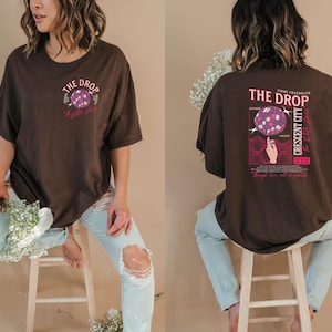 May include: A brown t-shirt with a graphic on the front and back. The front graphic says "The Drop" with a disco ball design. The back graphic says "The Drop Crescent City" with a disco ball design and a hand. The shirt is worn with light blue jeans.