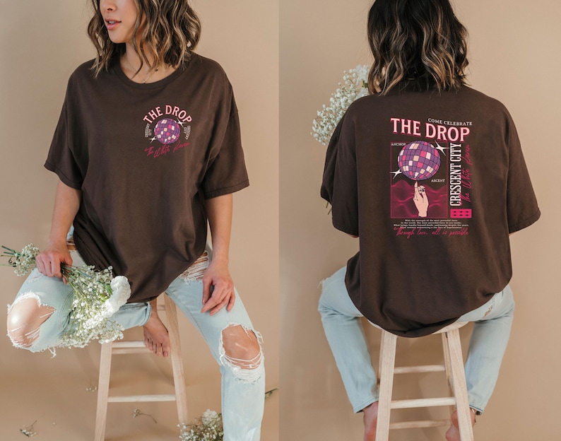 May include: A brown t-shirt with a graphic on the front and back. The front graphic says "The Drop" with a disco ball design. The back graphic says "The Drop Crescent City" with a disco ball design and a hand. The shirt is worn with light blue jeans.