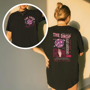 May include: Black t-shirt with a graphic on the front and back. The front graphic says "The Drop" with a disco ball design. The back graphic says "Come Celebrate The Drop" with a disco ball design and the words "Crescent City" and "The White Dawn".