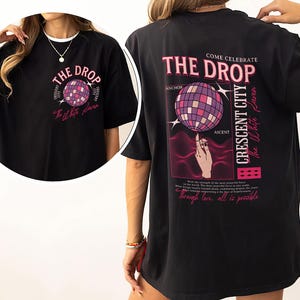 May include: Black t-shirt with a graphic design featuring the text "THE DROP" and a disco ball. The back of the shirt includes additional text: "COME CELEBRATE THE DROP CRESCENT CITY The White Paven".