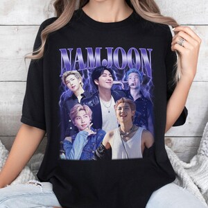 May include: Black t-shirt featuring a graphic of a group of people with the name "NAMJOON" in a purple, retro font. The graphic has a vintage aesthetic with a lightning bolt design in the background.