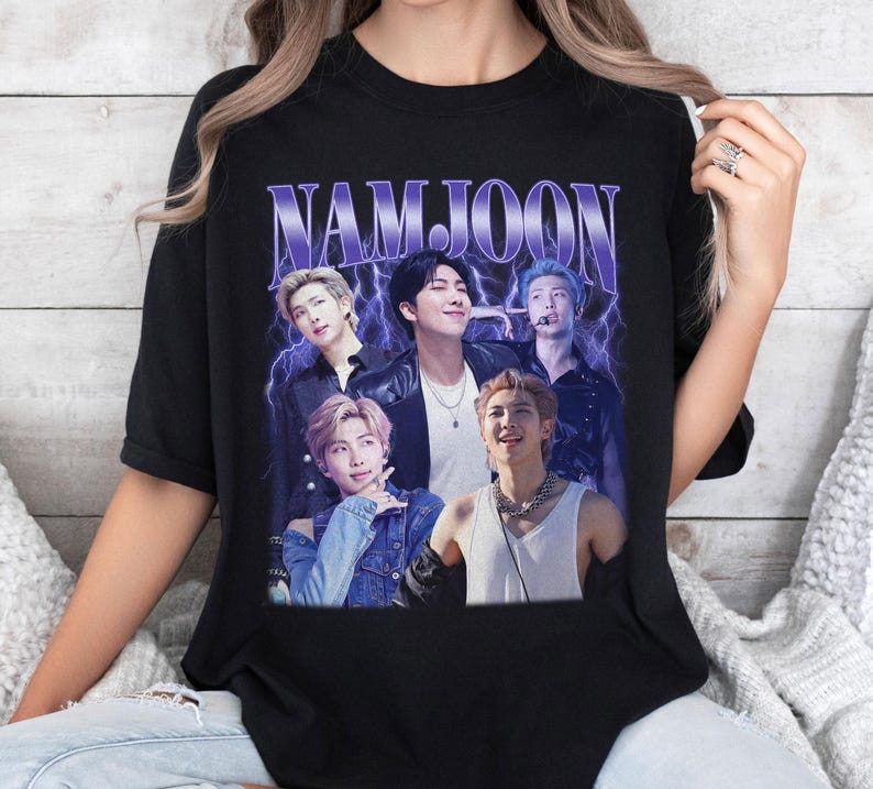 May include: Black t-shirt featuring a graphic of a group of people with the name "NAMJOON" in a purple, retro font. The graphic has a vintage aesthetic with a lightning bolt design in the background.