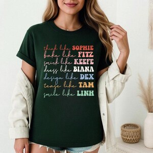 Puede incluir: Camiseta verde oscuro con texto colorido: "think like SOPHIE, bake like FITZ, smirk like KEEFE, dress like BIANA, design like DEX, tease like TAM, smile like LINH." El texto es de varios colores y fuentes. La camiseta tiene mangas cortas.