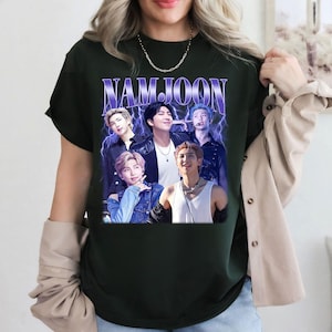 May include: Dark green t-shirt featuring a graphic of the word "NAMJOON" in purple and white, with a collage of five people. The design has a vintage, retro aesthetic with lightning bolt accents.