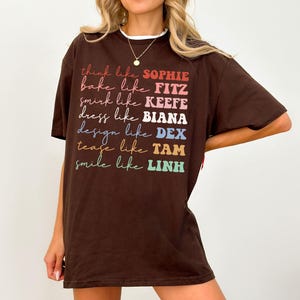 Puede incluir: Una camiseta marr&oacute;n con las frases "think like SOPHIE, bake like FITZ, smirk like KEEFE, dress like BIANA, design like DEX, tease like TAM, smile like LINH" impresas en varios colores. La camiseta tiene un cuello blanco.
